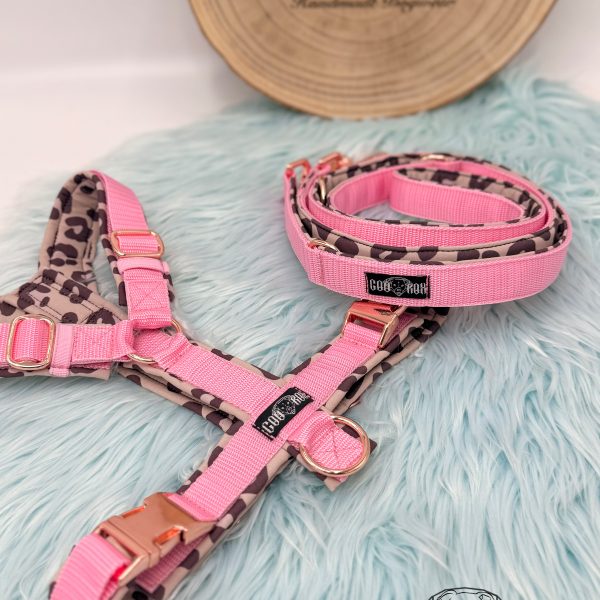 Handgemachtes Hundegeschirr mit passender Leine in Rosa mit rosa Leopardenmuster, Softshell gepolstert, roségoldener Hardware und Coorox-Label – fotografiert auf hellblauem Kunstfell vor gravierter Holzplatte.